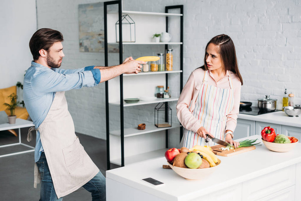girlfriend cutting vegetables and serious boyfriend having fun with banana gun in kitchen - Foto, immagini
