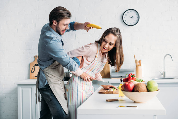 fidanzata taglio verdure e fidanzato divertirsi con banana pistola in cucina
 - Foto, immagini