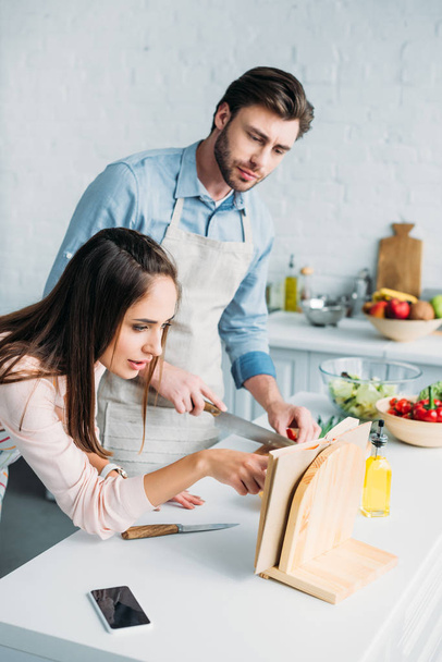 boyfriend cutting vegetables and girlfriend reading recipe in kitchen - Фото, изображение