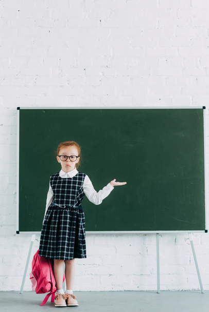 adorale little schoolgirl in eyeglasses holding pink school bag and showing blank chalkboard - Foto, Imagem