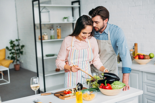 girlfriend cooking and mixing salad in kitchen and boyfriend hugging her - Photo, Image