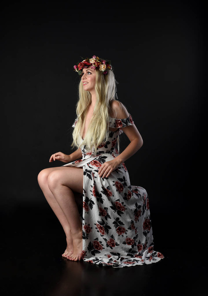 full length portrait of blonde girl wearing flower crown and floral dress, seated pose. isolated on black background. - Фото, изображение
