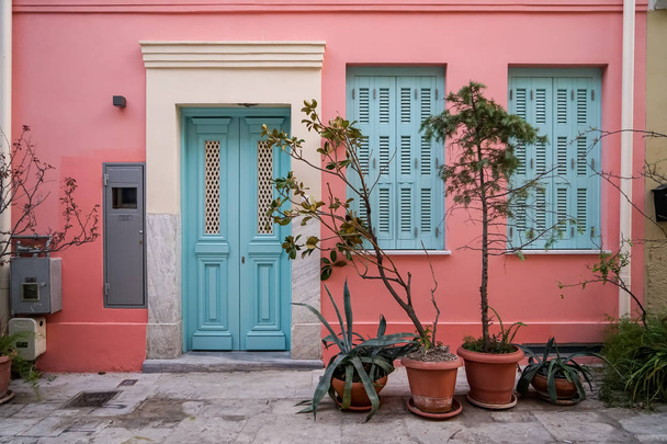 Escena de hermoso fondo de fachada de edificio urbano en pared de pintura de yeso rosa pastel, puerta de entrada azul claro y ventanas con macetas verdes, Atenas, Grecia
 - Foto, Imagen