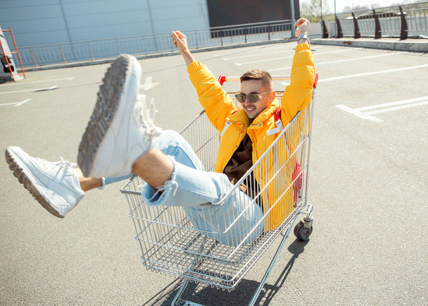 fashion guy in sunglasses and a yellow jacket jump in a cart from food in the supermarket parking - Photo, image
