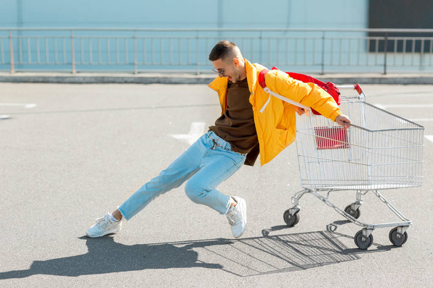 mode kerel in zonnebril en een gele jas springen in een kar van levensmiddelen in de supermarkt parking - Foto, afbeelding