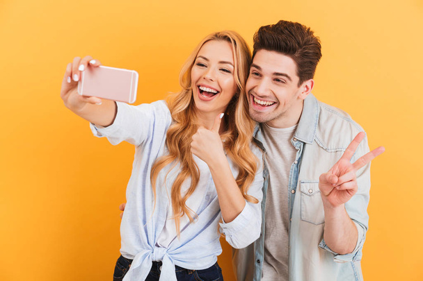Portrait of happy couple man and woman taking selfie photo on cell phone while gesturing at camera with fingers isolated over yellow background - Foto, imagen