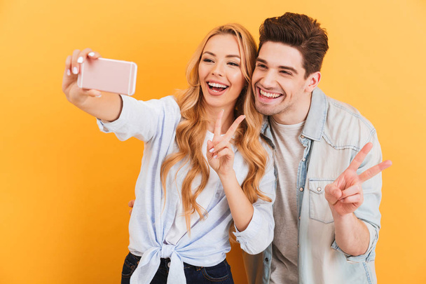 Portrait of young man and woman taking selfie photo on mobile phone while gesturing at camera victory sign isolated over yellow background - Photo, image