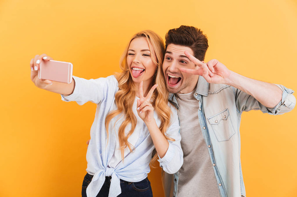 Portrait of funny man and woman taking selfie photo on mobile phone while gesturing at camera peace symbols isolated over yellow background - 写真・画像