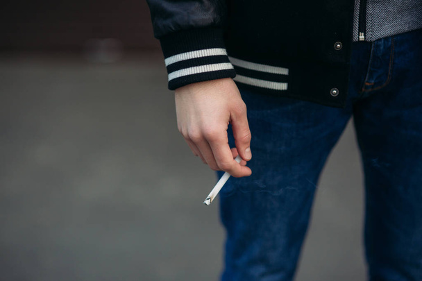 A young guy in a black jacket against a background of dark striped walls smoke cigarette - Foto, Imagen