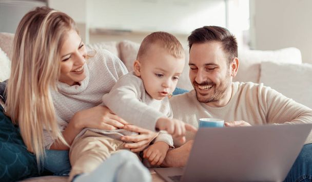 Χαμογελώντας γονείς και ο μικρός γιος που χρησιμοποιούν φορητό υπολογιστή στο σπίτι. - Φωτογραφία, εικόνα