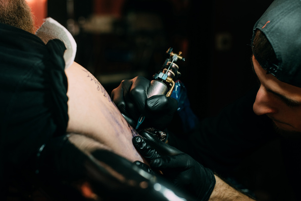 szelektív összpontosít a tetoválás művész a tetoválás gép működik a tetoválás a vállán a szalonban kesztyű - Fotó, kép