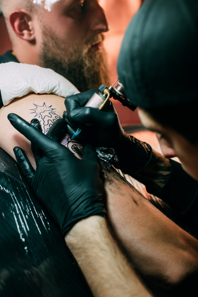 szelektív összpontosít a tetoválás művész a tetoválás gép működik a tetoválás a vállán a szalonban kesztyű - Fotó, kép