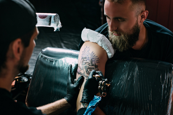 enfoque selectivo del artista del tatuaje en guantes con máquina de tatuaje trabajando en el tatuaje en el hombro en el salón
 - Foto, Imagen