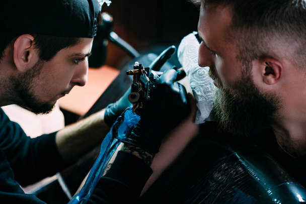 oldalnézetből a tetoválás művész a tetoválás gép működik a tetoválás a vállán a szalonban kesztyű - Fotó, kép