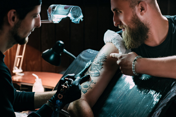 vista lateral del artista del tatuaje en guantes con máquina de tatuaje trabajando en el tatuaje en el hombro en el salón
 - Foto, Imagen