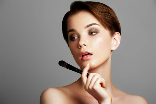 Beautiful young woman applying foundation on her face with a makeup brush isolated on gray background. Attractive young caucasian model. girl applying cosmetic powder on her face, skin care concept - Photo, Image