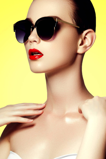 Μόδας γυαλιά ηλίου. Σέξι γυναίκα σε μαγιό με χρυσά γυαλιά ηλίου. Αίγλη βολή της ένα όμορφο μοντέλο σε λευκό μπικίνι και σκούρα γυαλιά ηλίου. Νεαρή κοπέλα ποζάρει με μαγιό άσπρο σε κίτρινο φόντο - Φωτογραφία, εικόνα