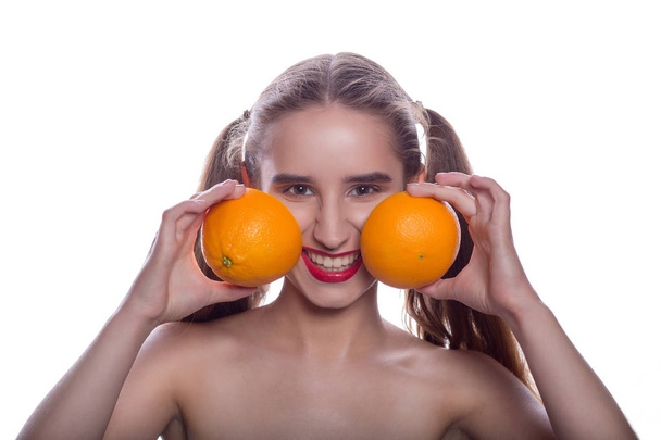 Pozytywne brunetka kobieta shirtless trzymając kawałki pomarańczy w pobliżu jej policzki, stojąc na białym tle - Zdjęcie, obraz