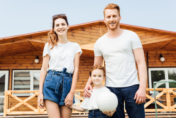 beautiful young family with volleyball ball standing in front of wooden cottage - Foto, immagini