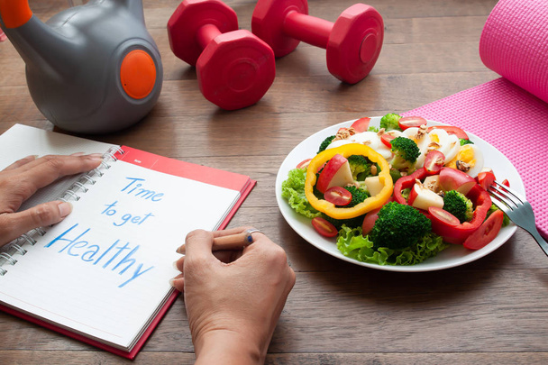 Woman writing on diary book with salad and fitness equipments on table. Dieting concept - Photo, image