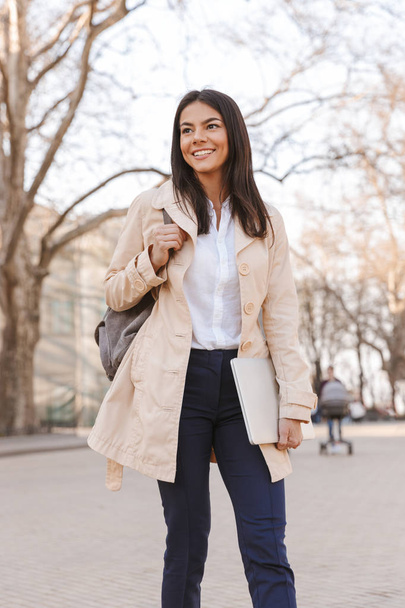 Cheerful young woman dressed in autumn coat carrying laptop while walking on a city street - Photo, image