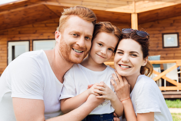 close-up shot of beautiful young family embracing in front of wooden cottage and looking at camera - Foto, afbeelding