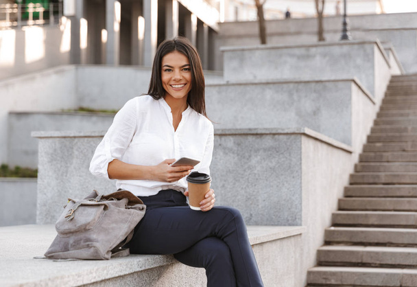 Happy business woman sitting outdoors with cup of coffee and smartphone while looking at the camera - Photo, Image