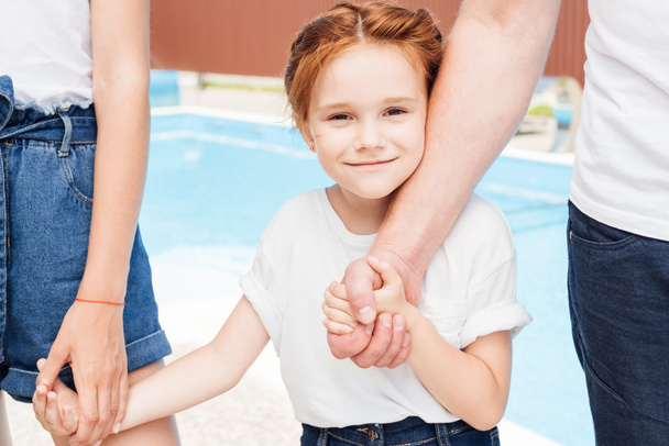 happy little girl holding hands of parents and looking at camera in front of swimming pool - Foto, Imagem