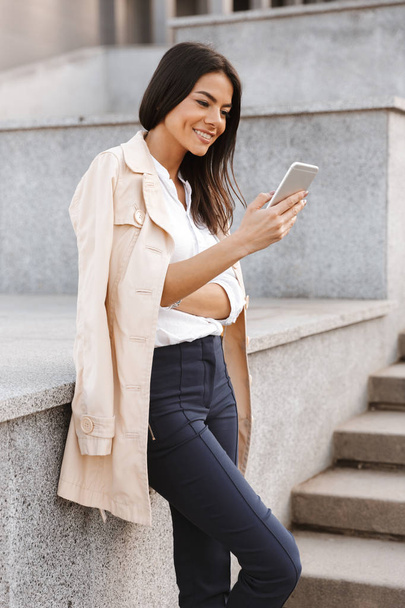 Smiling young woman using mobile phone while standing on a stairs outdoors - Photo, image