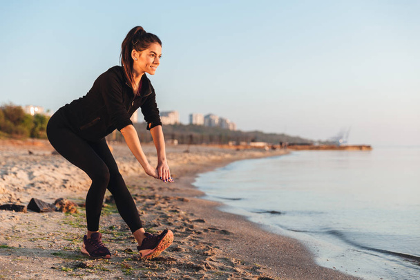 Happy νεαρό αθλήτρια που κάνει το τέντωμα ασκήσεις στην παραλία το πρωί - Φωτογραφία, εικόνα