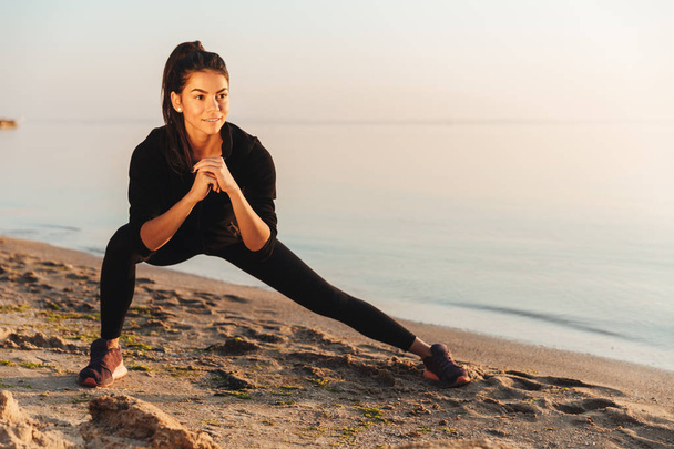 Smiling young sportswoman doing stretching exercises at the seaside in the morning - Фото, изображение