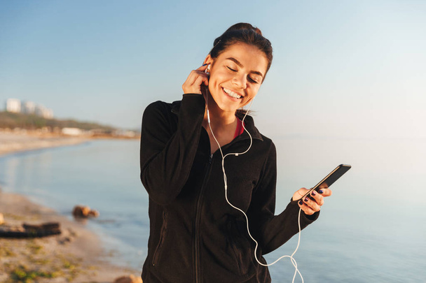 Satisfied young sportswoman listening to music with earphones and mobile phone while standing at the beach - Фото, изображение