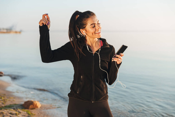 Delighted young sportswoman listening to music with earphones and mobile phone while standing at the beach - Foto, Imagem