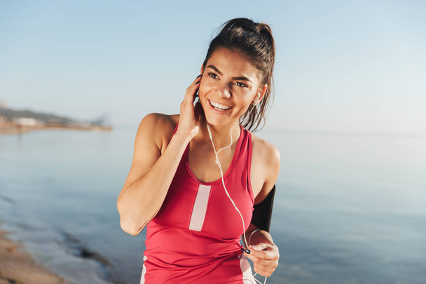 Smiling sports woman listening music by smartphone and looking away near the sea outdoors - Foto, afbeelding