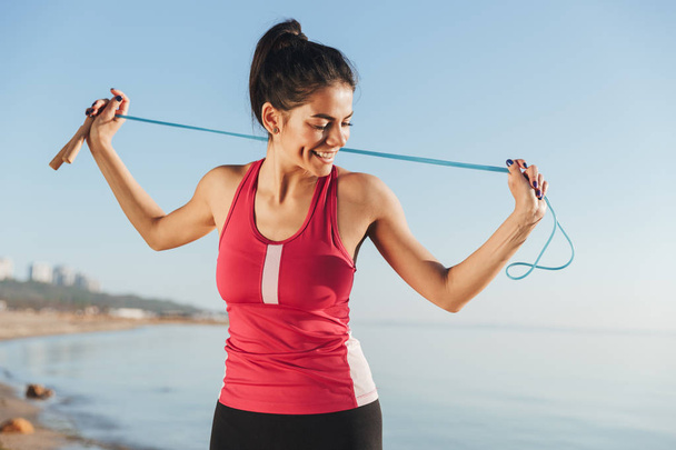 Pleased sports woman posing with jumping rope and looking away near the sea outdoors - Photo, image