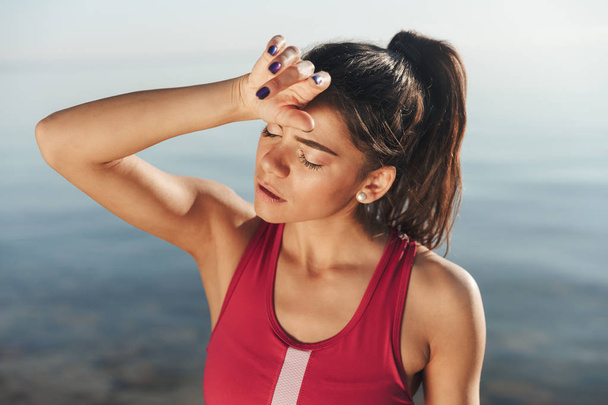 Exhausted sportswoman wiping her forehead after jogging while standing at the beach - Foto, afbeelding