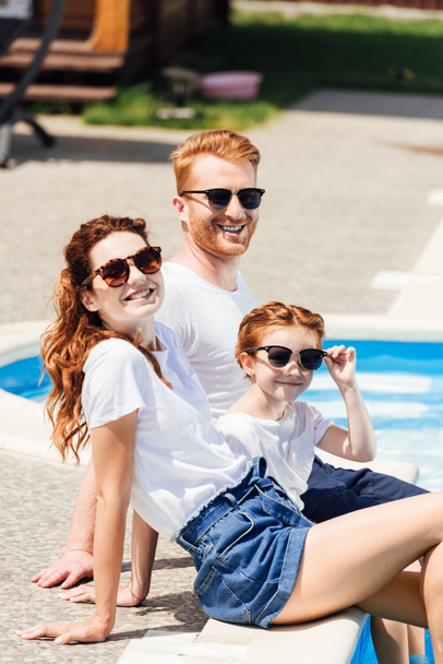 beautiful young family in white t-shirts and sunglasses sitting on poolside together and looking at camera - Foto, immagini