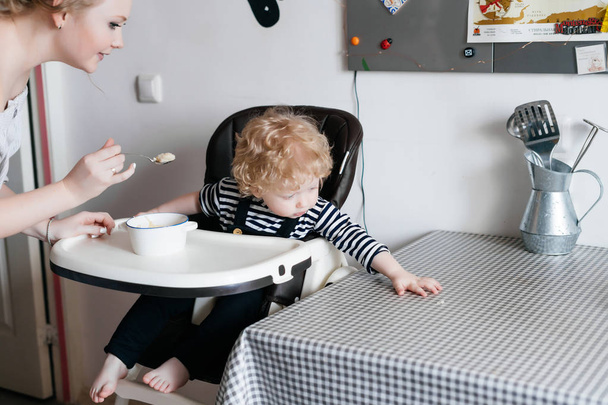 a caring young mother feeds her curly little son with a useful tasty soup in the kitchen - Fotoğraf, Görsel