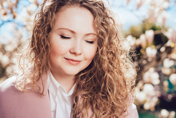 beautiful curly young woman posing in park next to blooming magnolia, enjoying rest and smiling - Фото, зображення