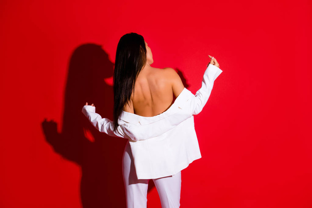 Rear view portrait of nude sensual girl taking off jacket showing naked shoulders isolated on vivid red background. Striptease concept - Photo, image