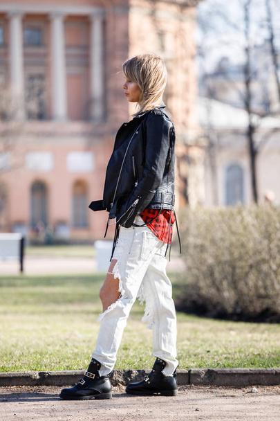 Outdoor portrait of beauty blond woman wearing black leather jacket, grey t-shirt and torn jeans standing in park, blurred city background  - Fotografie, Obrázek
