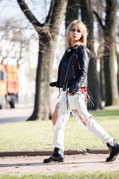 Outdoor portrait of beauty blond woman wearing black leather jacket, grey t-shirt and torn jeans standing in park, blurred city background  - 写真・画像