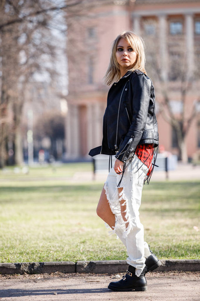Outdoor portrait of beauty blond woman wearing black leather jacket, grey t-shirt and torn jeans standing in park, blurred city background  - Foto, Imagen