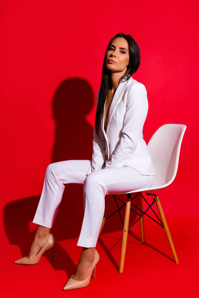 Full size portrait of stunning luxury woman sitting on stool wearing white suit looking at camera isolated on bright red background - Фото, изображение