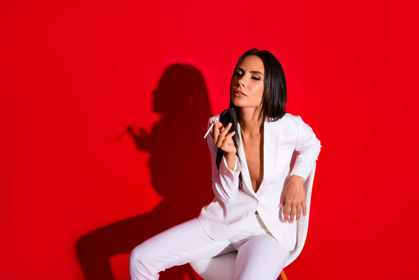 Portrait of pleased delightful woman enjoying smoking cigarette wearing white suit with tempting decolette isolated on bright red background - Fotó, kép