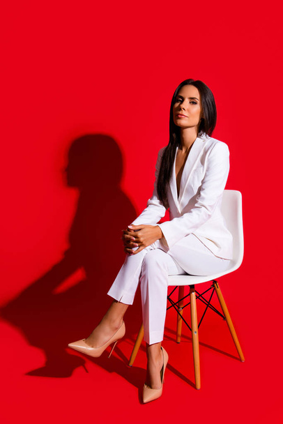 Photoshooting studio concept. Vertical portrait of proud arrogant woman sitting on chair leg by foot looking at camera isolated on vivid red background - Photo, Image