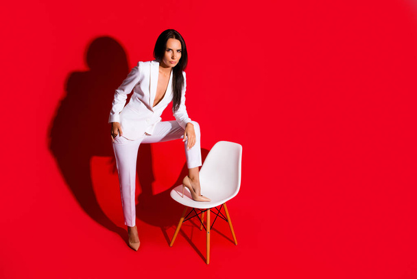 Portrait of skinny confident woman putting leg on chair looking at camera wearing white suit with sexual decollete isolated on vivid red background. Photoshooting studio concept - Foto, afbeelding