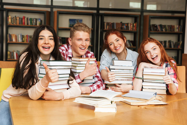 Skupina veselých teenagerů sedí u stolu knihovny s velkým stohy knih a ukazuje palec - Fotografie, Obrázek