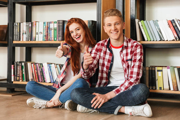 Sonriente pareja de adolescentes sentados en un piso en la estantería de una biblioteca y mostrando los pulgares hacia arriba
 - Foto, Imagen