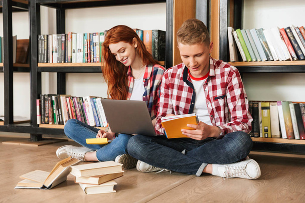 Smiling teenage couple sitting on a floor at the bookshelf in a library and doing homework with laptop computer - 写真・画像